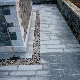 Kamienny taras z szarej kostki brukowej i obrzeżem z kamienia naturalnego, oddzielony od ściany budynku pasem z jasnych otoczaków, z miotłą opartą o ścianę.