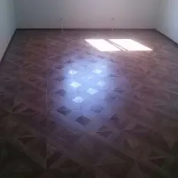 Nowo ułożona, ciemnobrązowa podłoga z paneli w geometryczny wzór, oświetlona przez promienie słoneczne wpadające przez okno, widoczne białe ściany i rury grzewcze.