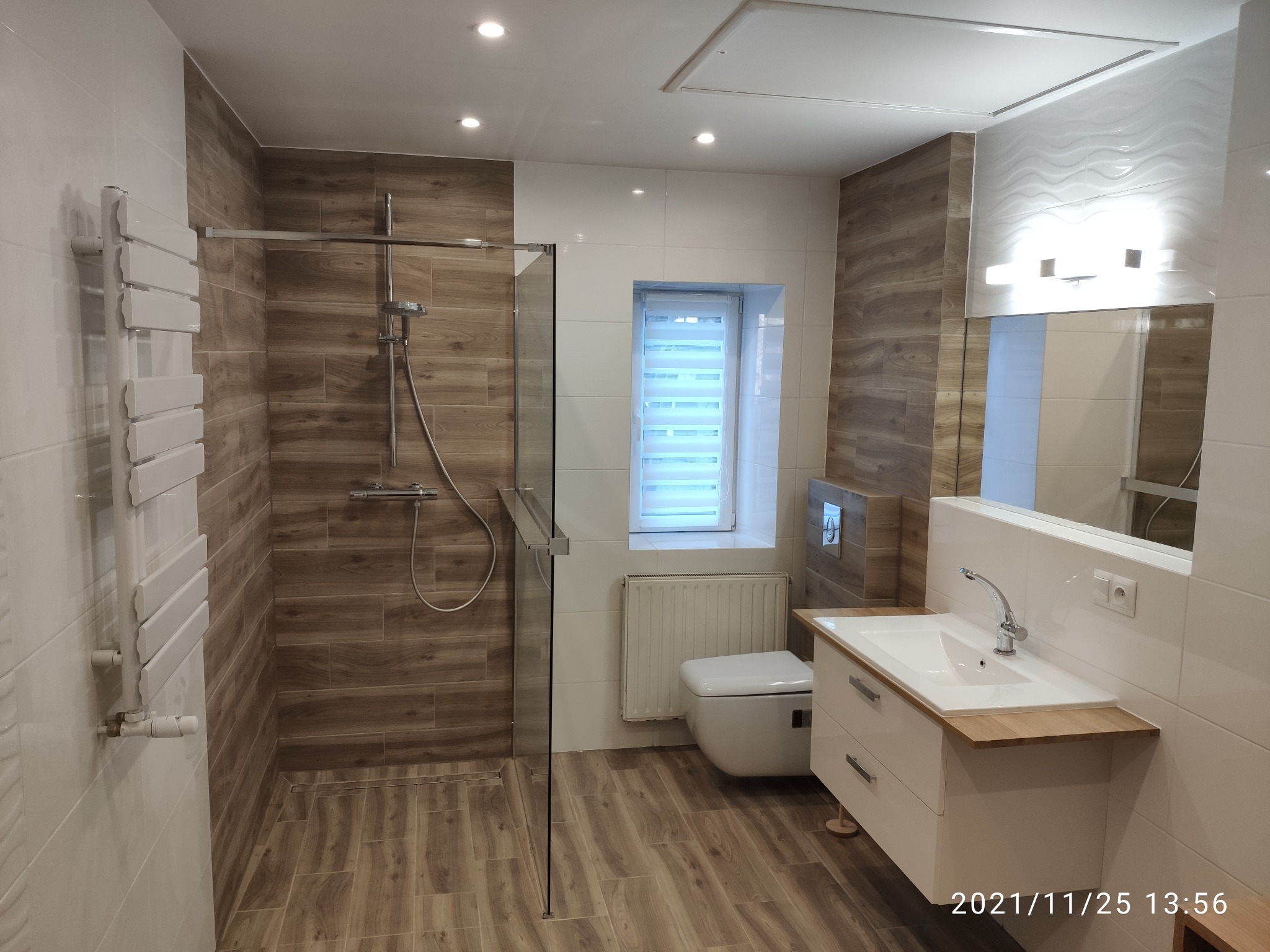 Nowoczesna łazienka z kabiną prysznicową typu walk-in, grzejnikiem drabinkowym, umywalką z szafką i toaletą w zabudowie, wykończona płytkami imitującymi drewno i białymi płytkami.