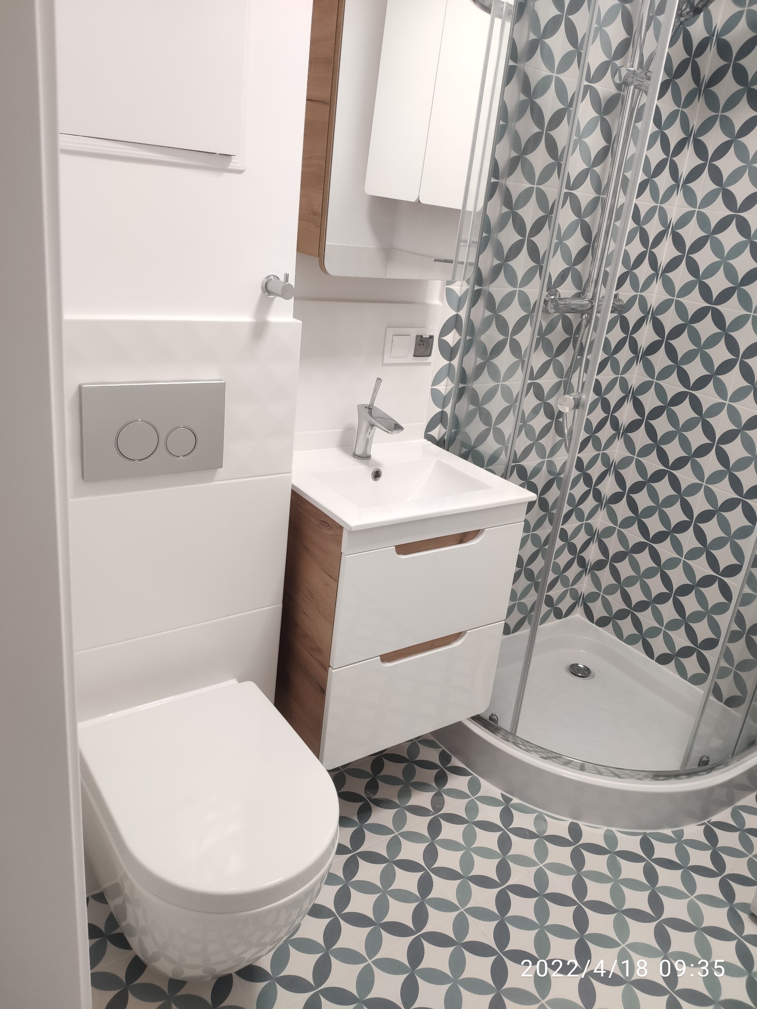 Nowoczesna, wykończona łazienka w Krakowie z kabiną prysznicową, umywalką z szafką w kolorze białym i drewnopodobnym, podwieszaną toaletą oraz wzorzystymi płytkami podłogowymi i ściennymi...