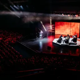 Szeroki kadr na scenę podczas konferencji z prelegentem przy mównicy i panelem dyskusyjnym, oświetloną reflektorami i z publicznością w tle.