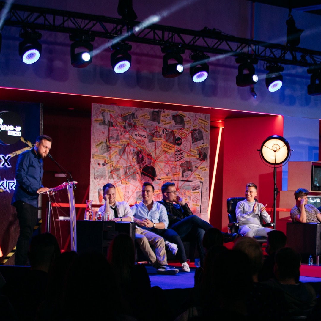 Konferencja: Mężczyzna w niebieskiej koszuli przemawia przy mównicy na scenie oświetlonej reflektorami, za nim siedzi panel gości na tle mapy powiązań.