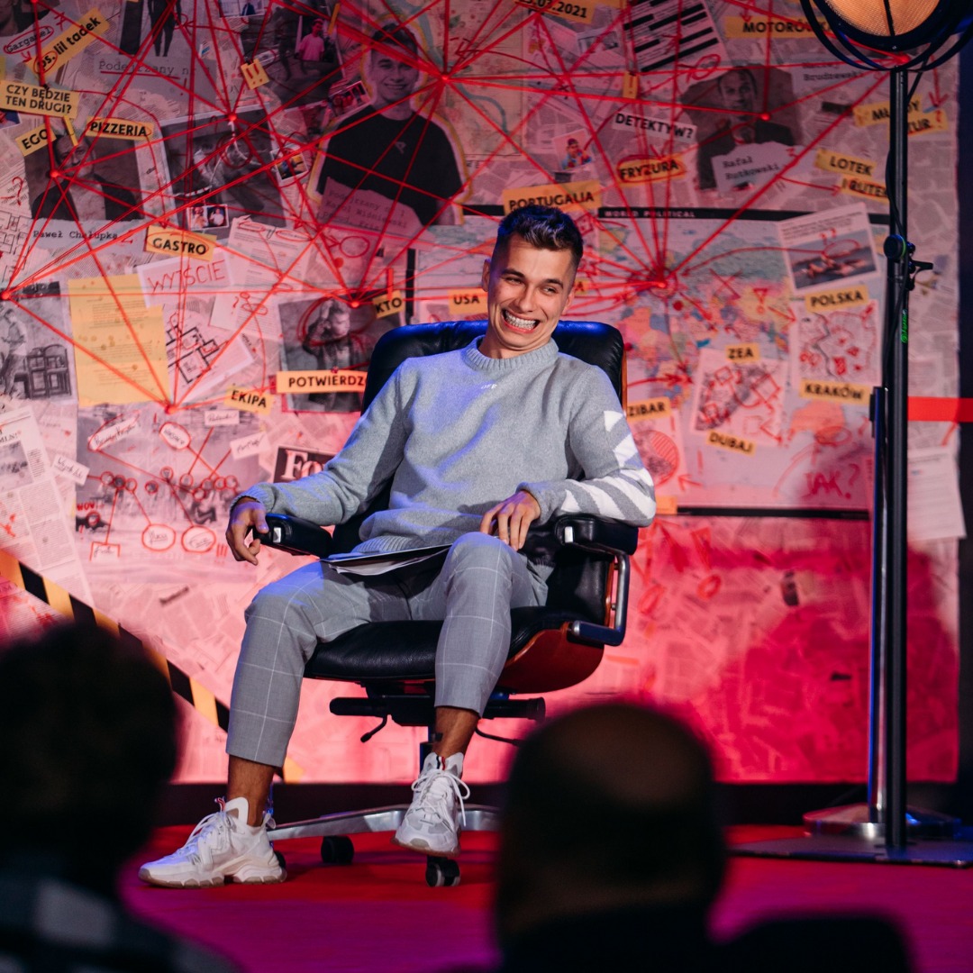 Uśmiechnięty mężczyzna w szarym swetrze siedzi na czarnym krześle na tle ściany z mapami, zdjęciami i czerwonymi nitkami, przypominającej tablicę detektywistyczną.