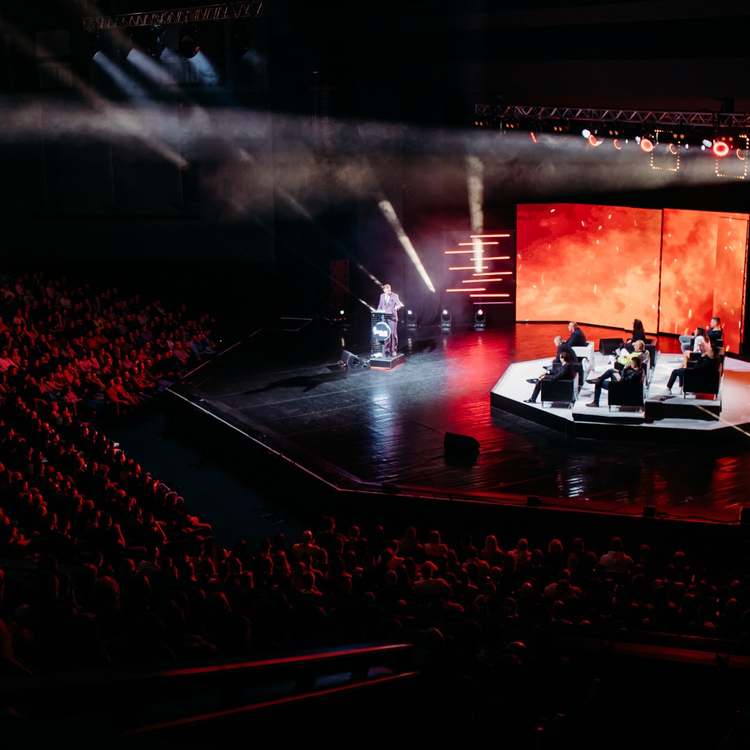 Szeroki kadr na scenę podczas konferencji z prelegentem przy mównicy i panelem dyskusyjnym, oświetloną reflektorami i z publicznością w tle.