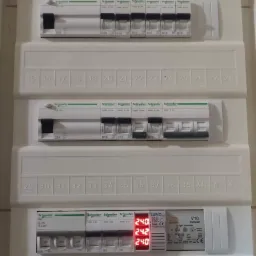 Rozdzielnica elektryczna z zainstalowanymi bezpiecznikami i modułem pomiarowym LUNC V10, wyświetlającym aktualne napięcie 240-242V, zamontowana na ścianie.