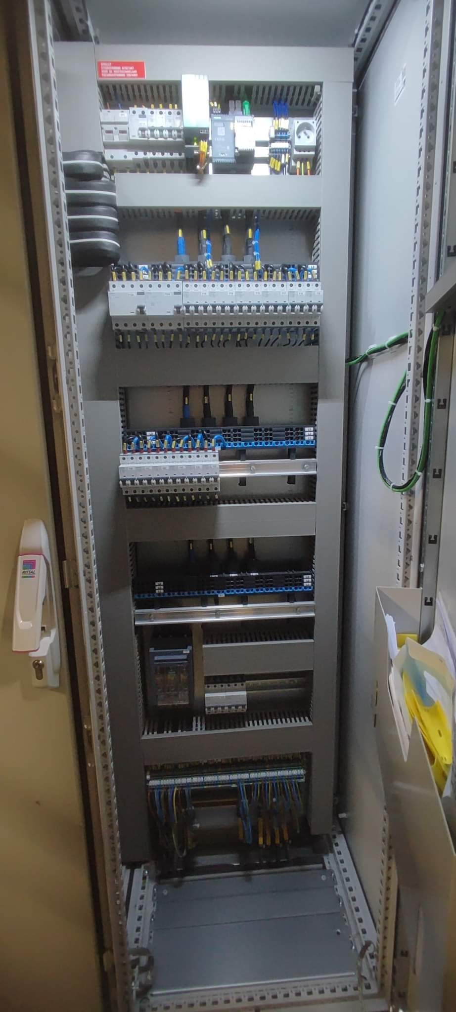 Otwarta szafa sterownicza z widocznymi zabezpieczeniami, przekaźnikami i okablowaniem, widok z boku, z elementami oznaczonymi etykietami.