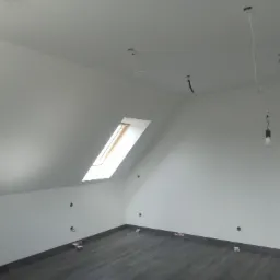 Poddasze z oknem dachowym, szare panele podłogowe częściowo ułożone, białe ściany z widocznymi instalacjami elektrycznymi i wystającymi kablami, żarówka na kablu.
