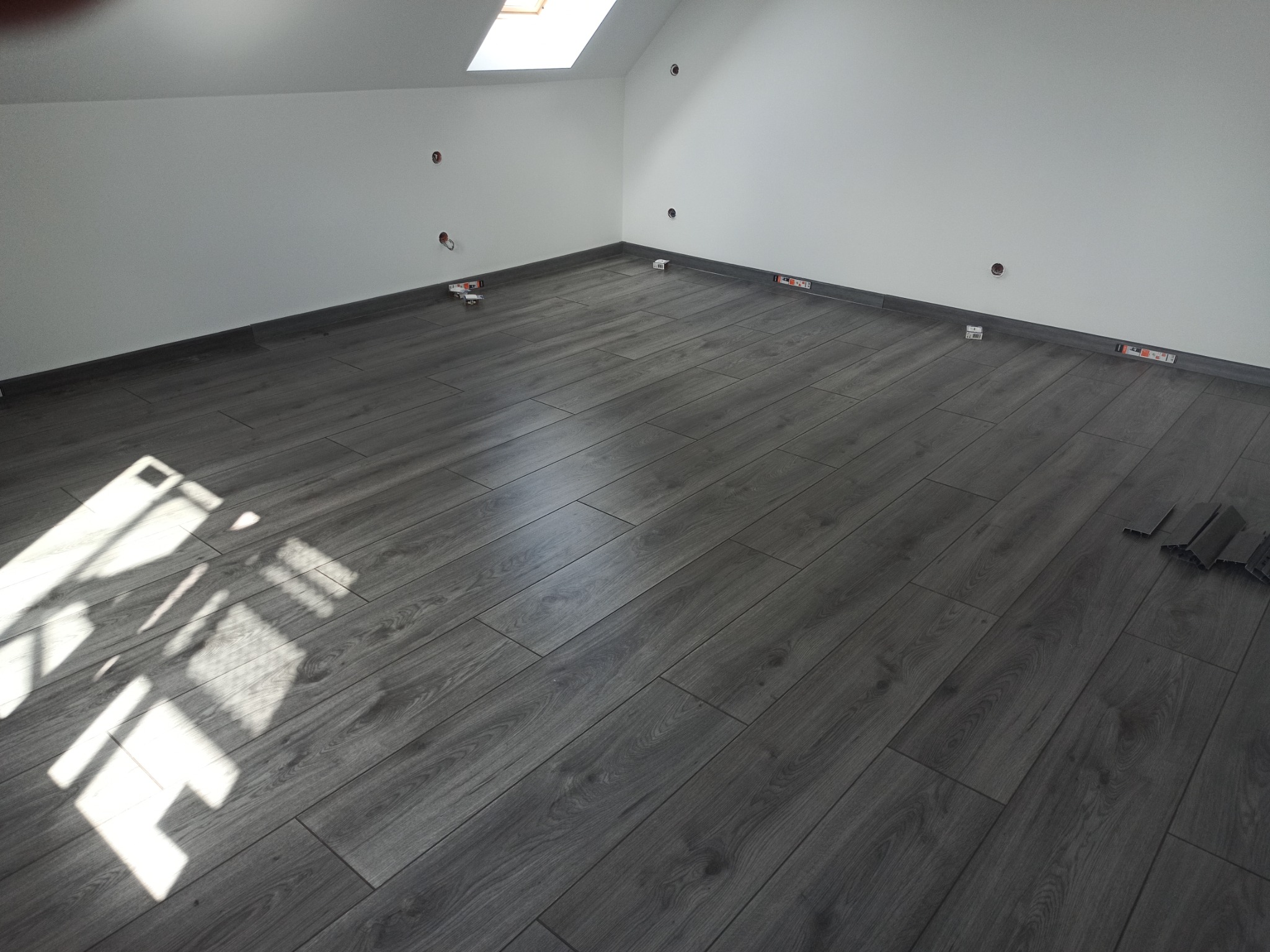 Ciemnoszare panele podłogowe ułożone w pokoju na poddaszu ze świetlikiem, widoczne listwy przypodłogowe i uchwyty montażowe, fragmenty paneli pod ścianą.