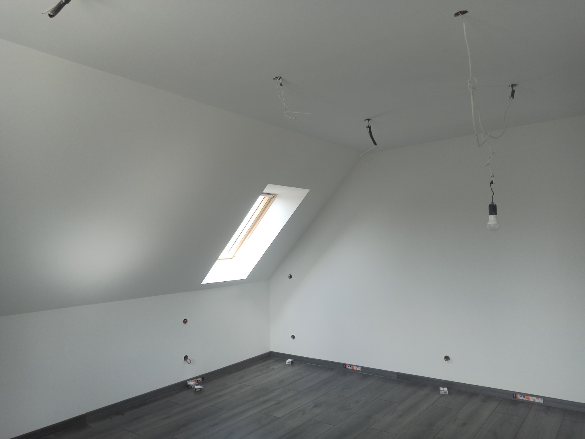 Poddasze z oknem dachowym, szare panele podłogowe częściowo ułożone, białe ściany z widocznymi instalacjami elektrycznymi i wystającymi kablami, żarówka na kablu.