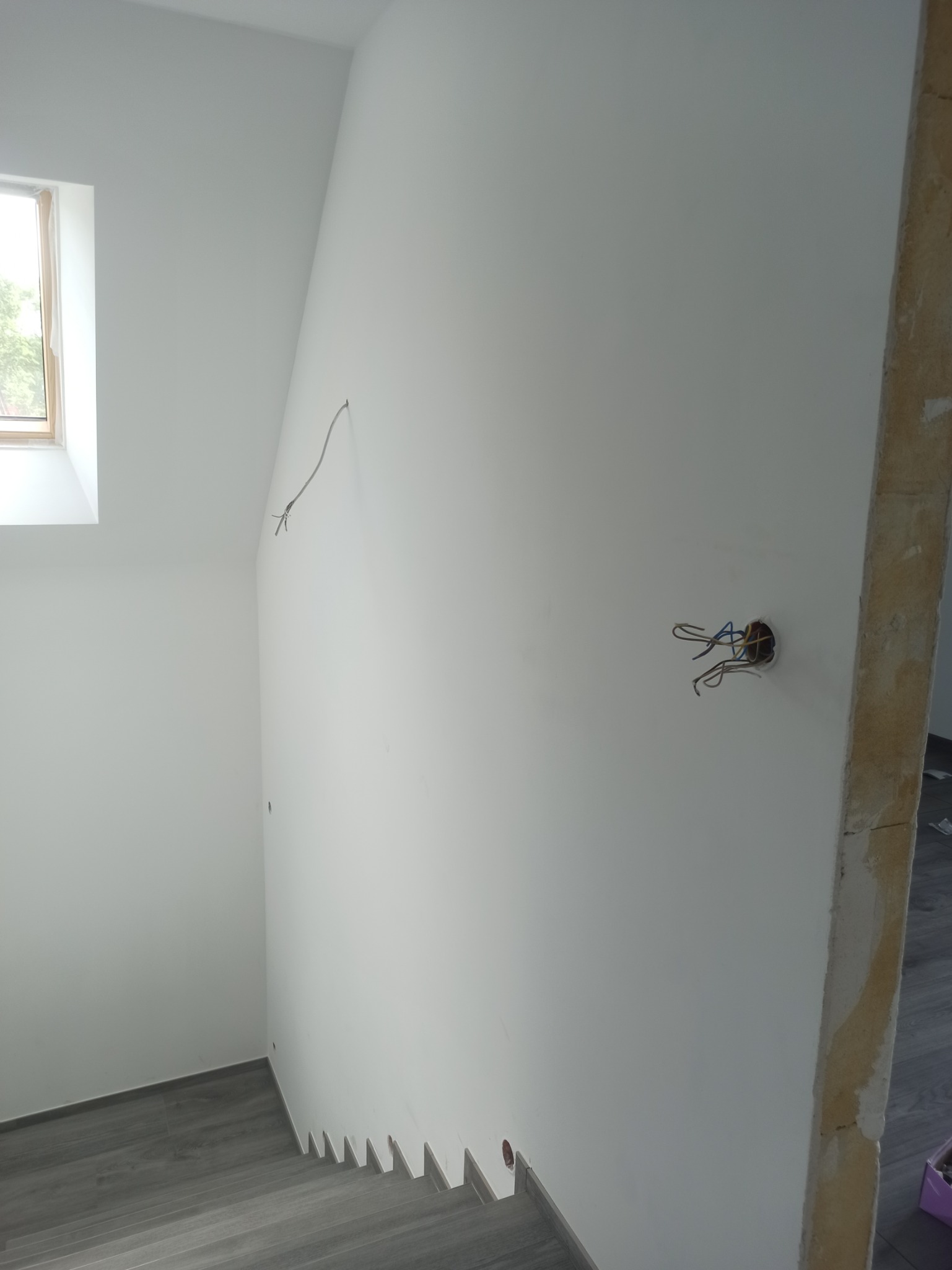 Wnętrze w trakcie prac wykończeniowych: białe ściany, szara podłoga imitująca drewno, widoczne przewody elektryczne wystające ze ściany, małe okno dachowe.