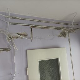 Michał Borowski MRenovations - Instalacja elektryczna w trakcie wykonywania w Błoniu: widoczne kable prowadzone w bruzdach na fioletowej ścianie, skrzynka z bezpiecznikami i fragment ościeżnicy drzwiowej.