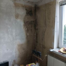 Michał Borowski MRenovations - Pokój w trakcie renowacji: ściany z widocznymi śladami wilgoci i starej farby, przygotowane do dalszych prac, widoczny worek z zaprawą, narzędzia i lampa oświetleniowa.