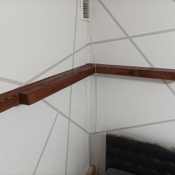 Michał Borowski MRenovations - Zamontowana, ciemnobrązowa drewniana półka narożna na białej ścianie z szarymi geometrycznymi wzorami, widoczny fragment łóżka i kratka wentylacyjna.