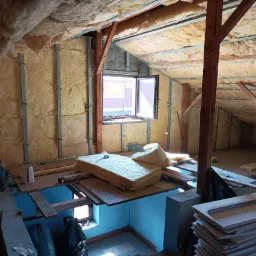 Poddasze w trakcie ocieplania: widoczne wełna mineralna, metalowe profile, drewniane belki konstrukcyjne, otwarte okno i stos płyt gipsowo-kartonowych.