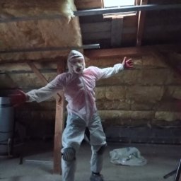 Michał Borowski MRenovations - Osoba w kombinezonie ochronnym i masce, z rozłożonymi ramionami, stoi na poddaszu z widoczną izolacją z wełny mineralnej i oknem dachowym.