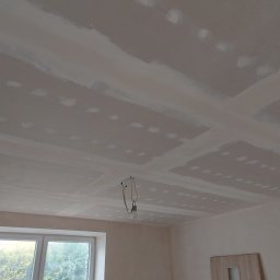 Michał Borowski MRenovations - Sufit w trakcie prac wykończeniowych: widoczne płyty gipsowo-kartonowe ze świeżo nałożonymi łatami, przygotowane do dalszej obróbki, wisząca żarówka na kablu, okno w tle.