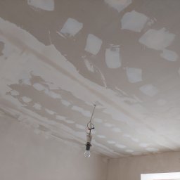 Michał Borowski MRenovations - Sufit w trakcie prac wykończeniowych, widoczne nałożone warstwy szpachli i gładzi, surowa żarówka zwisa na kablu.