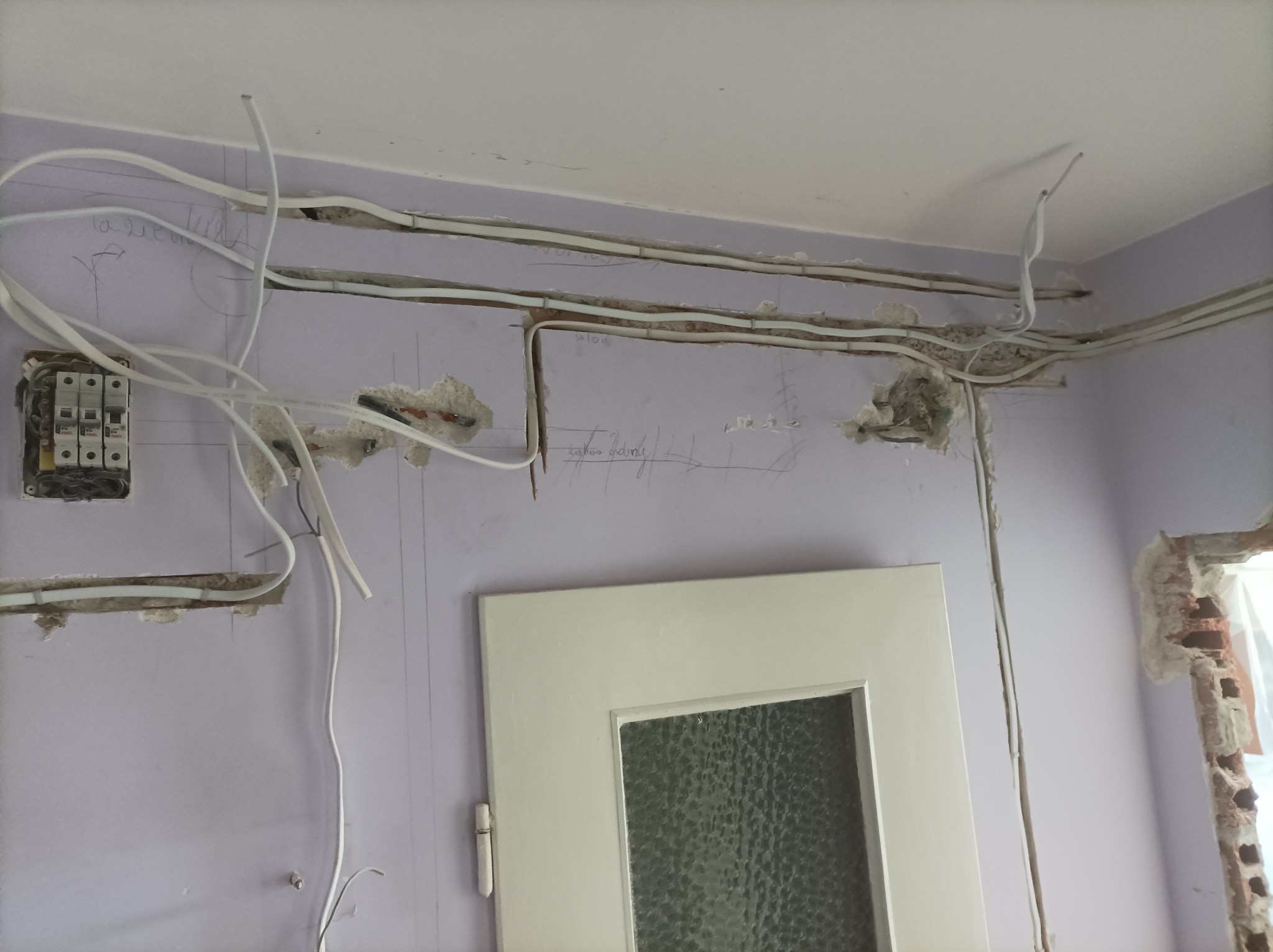 Instalacja elektryczna w trakcie wykonywania w Błoniu: widoczne kable prowadzone w bruzdach na fioletowej ścianie, skrzynka z bezpiecznikami i fragment ościeżnicy drzwiowej.