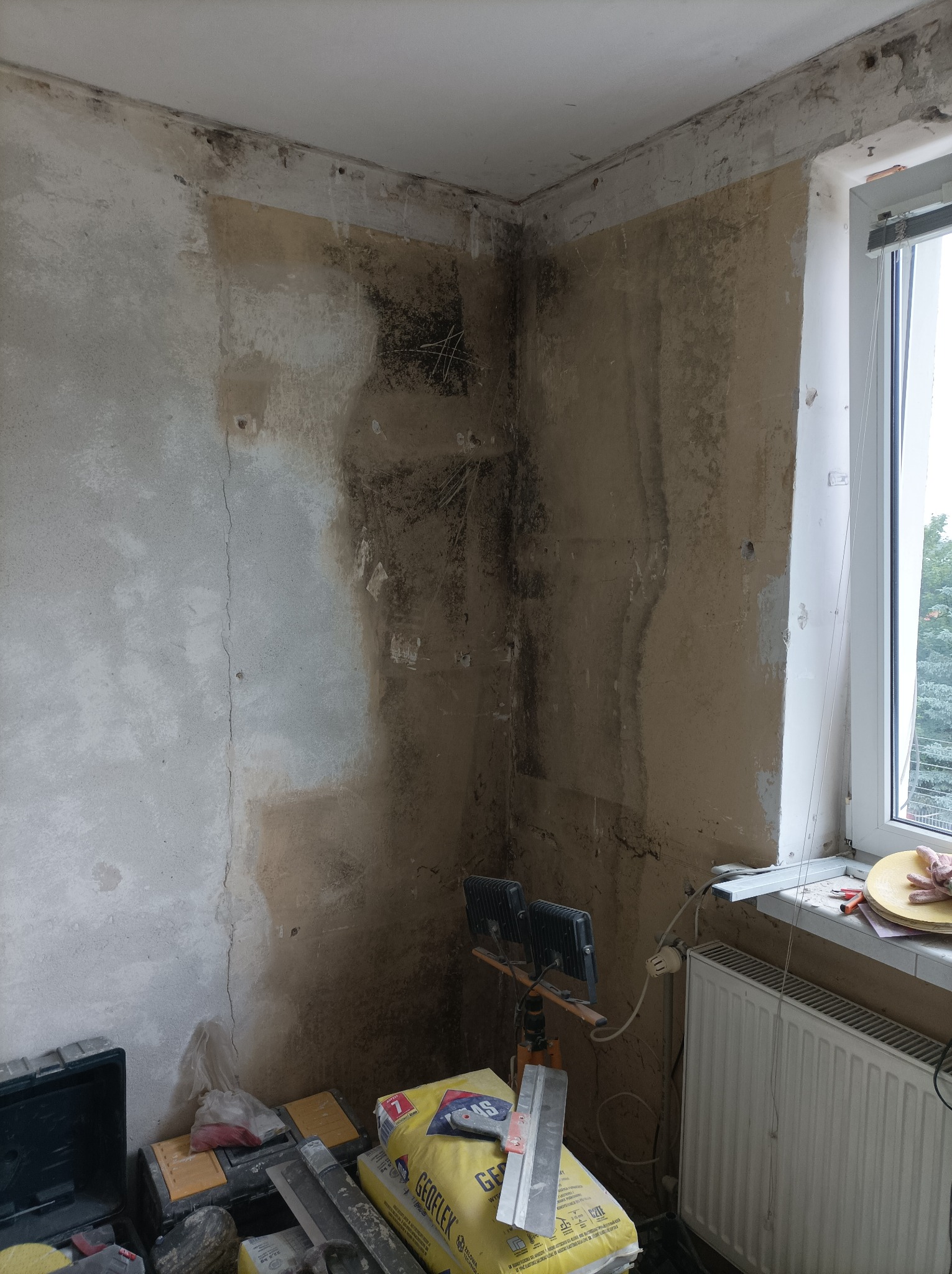 Pokój w trakcie renowacji: ściany z widocznymi śladami wilgoci i starej farby, przygotowane do dalszych prac, widoczny worek z zaprawą, narzędzia i lampa oświetleniowa.