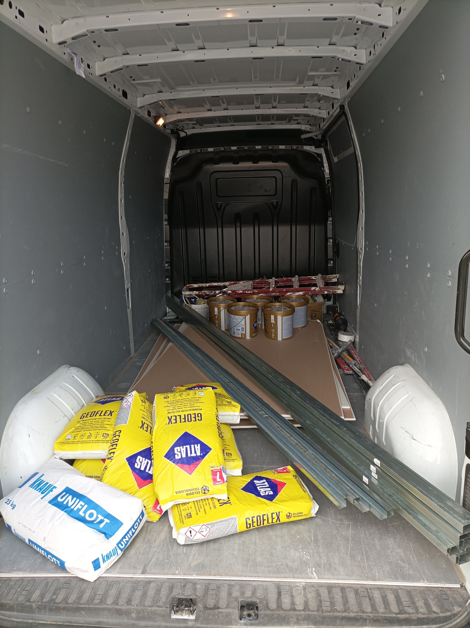 Wnętrze furgonetki załadowane materiałami budowlanymi: worki z klejem Atlas Geoflex i Knauf Uniflott, profile metalowe, płyty gipsowo-kartonowe oraz puszki z farbą.