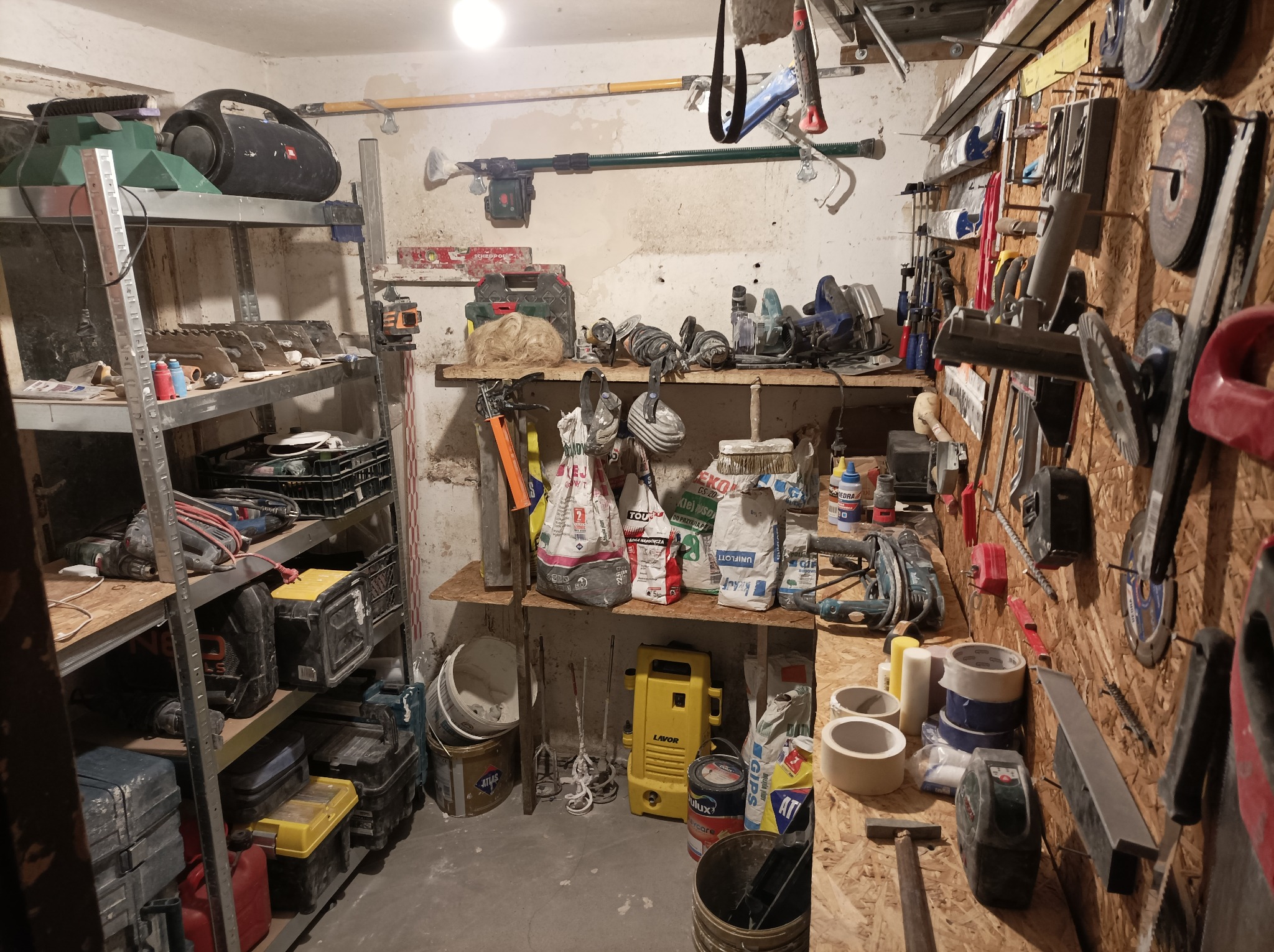 Warsztat z narzędziami i materiałami budowlanymi: półki z elektronarzędziami, worki z zaprawą, pędzle, szpachle i inne akcesoria do prac wykończeniowych.