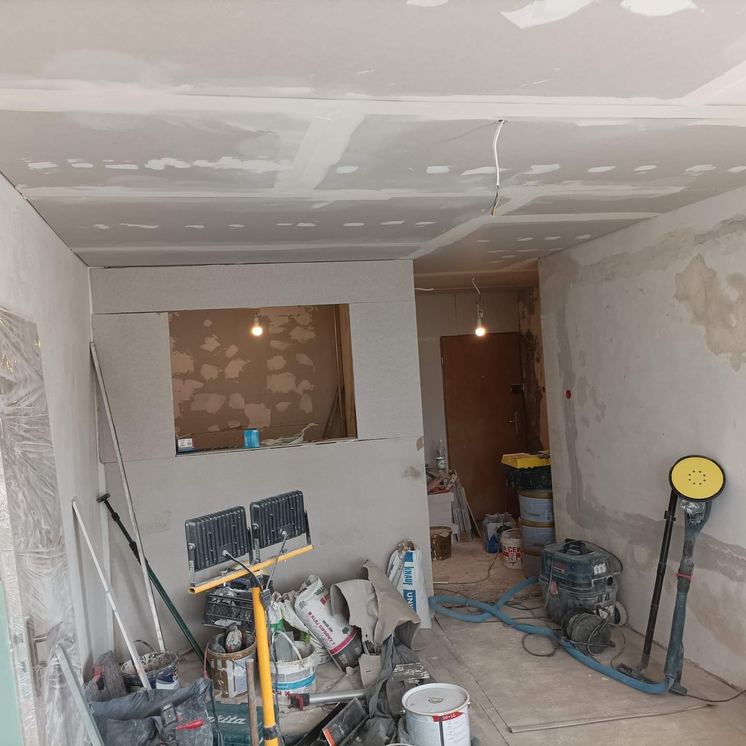 Wnętrze remontowanego pokoju z widocznymi ścianami i sufitem po szpachlowaniu, z pozostawionymi narzędziami i workami z materiałami budowlanymi, dwa reflektory oświetlają ścianę z otworem okiennym.