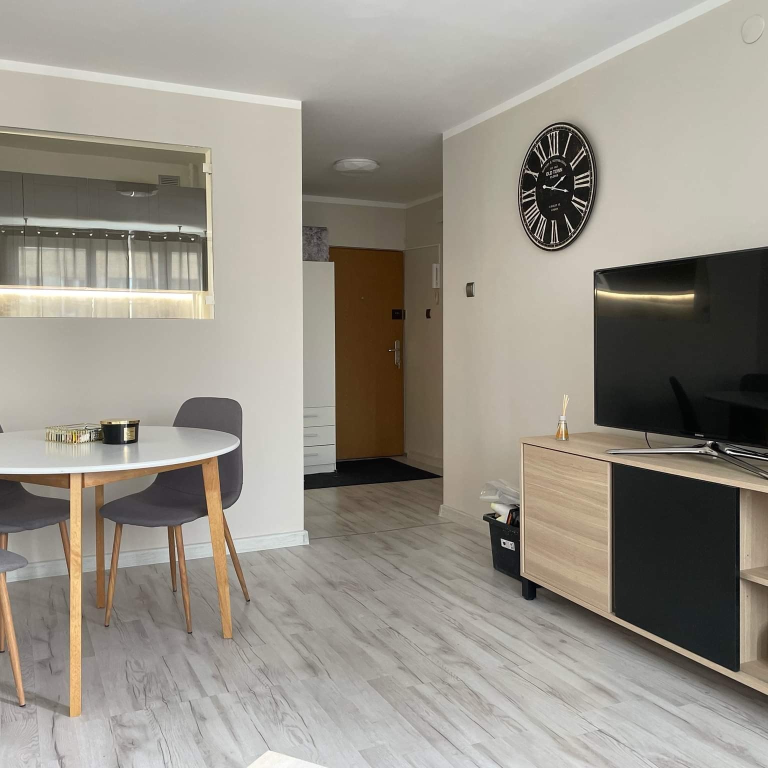 Jasne wnętrze mieszkania z okrągłym stołem, szarymi krzesłami, telewizorem na szafce RTV i dużym zegarem na ścianie, widok na przedpokój z drzwiami wejściowymi.