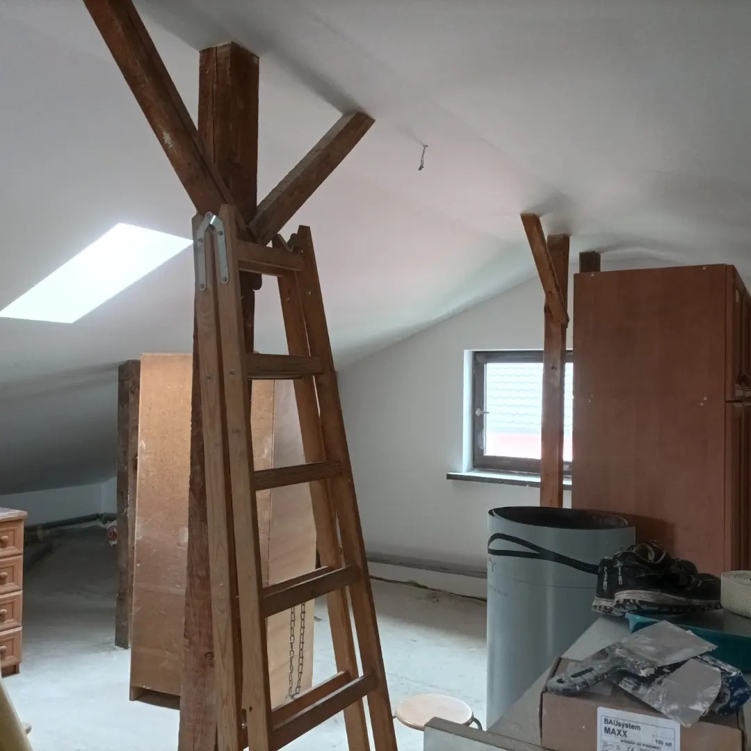Poddasze w trakcie prac wykończeniowych: drewniana drabina oparta o belkę, widoczne narzędzia, wiadro i resztki materiałów budowlanych, okno dachowe, ściany przygotowane do malowania.