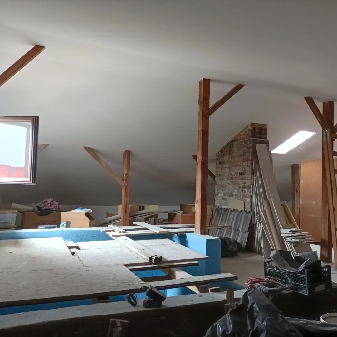 Poddasze w trakcie adaptacji: widoczne drewniane konstrukcje nośne, fragment murowanego komina, płyty karton-gips i materiały budowlane, niebieska konstrukcja pod podłogę, okno dachowe wpadające...