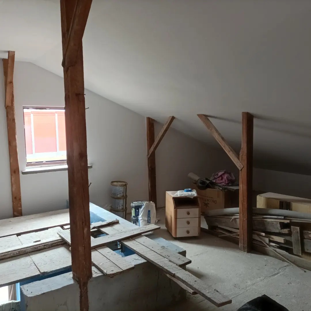 Poddasze w trakcie prac wykończeniowych: widoczne drewniane belki konstrukcyjne, okno, częściowo pomalowane ściany, płyty OSB, puszki z farbą i inne materiały budowlane, ukazujące etap...
