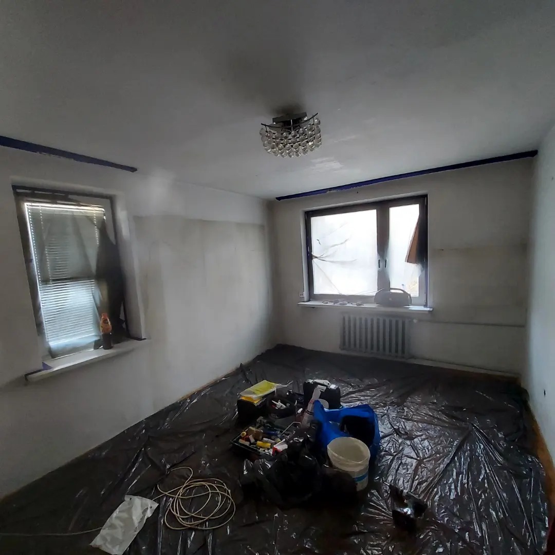 Pokój w trakcie przygotowania do prac malarskich: zabezpieczona folią podłoga, taśma malarska przy suficie, częściowo zagruntowane ściany, widoczne narzędzia i akcesoria malarskie.