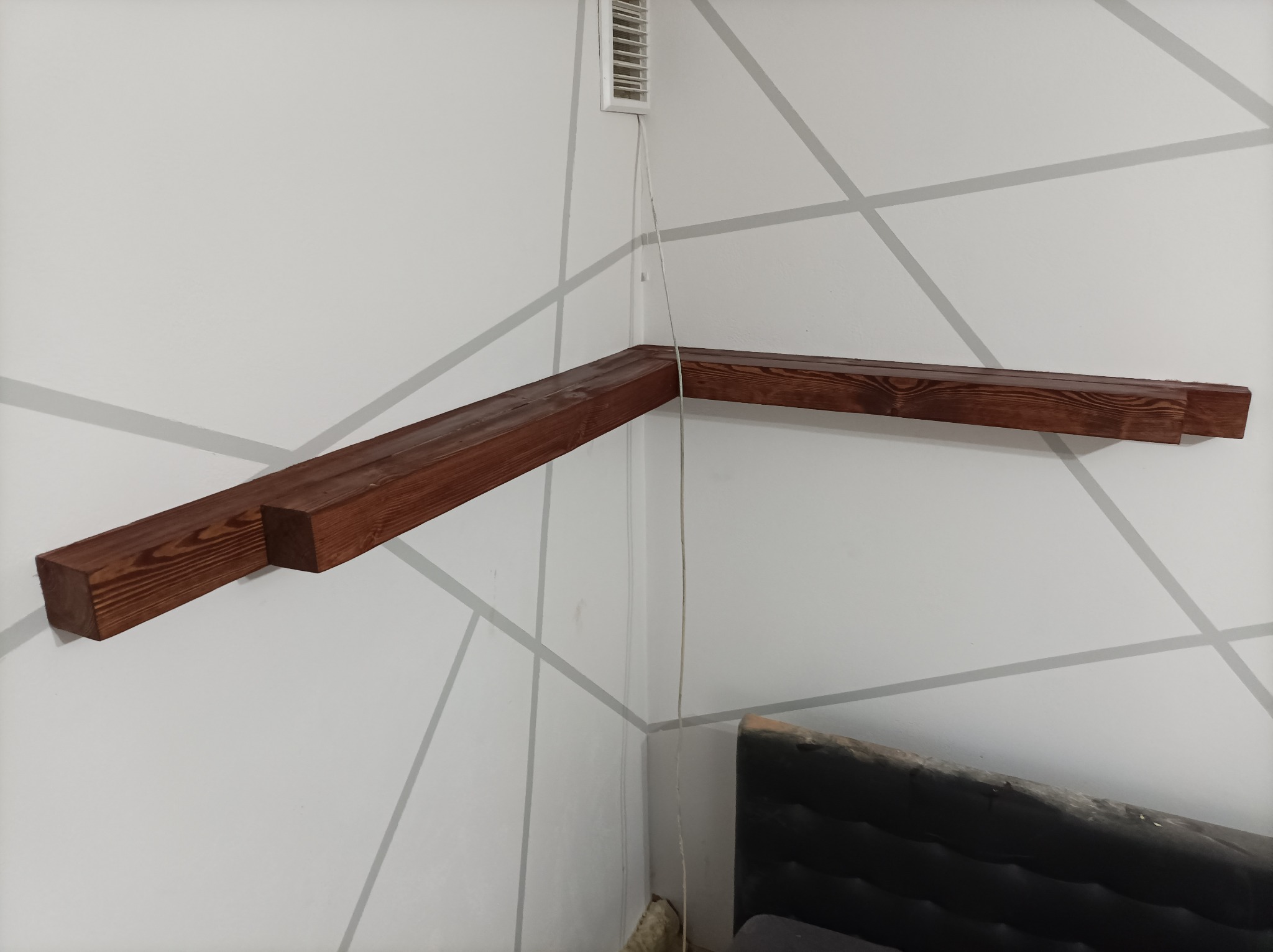 Zamontowana, ciemnobrązowa drewniana półka narożna na białej ścianie z szarymi geometrycznymi wzorami, widoczny fragment łóżka i kratka wentylacyjna.