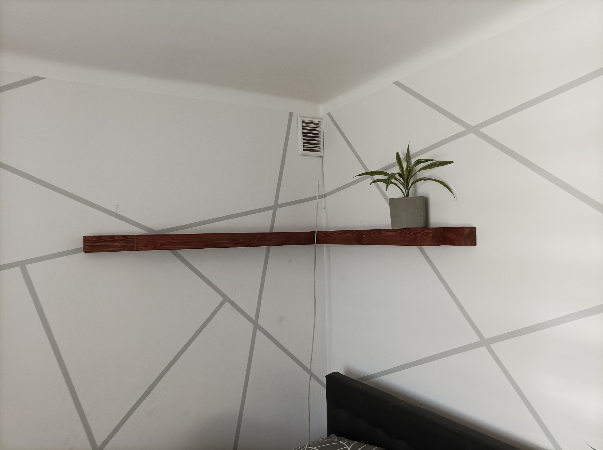 Półka z ciemnego drewna zamontowana w rogu pokoju na tle białej ściany z szarymi geometrycznymi wzorami, na półce stoi szara doniczka z zieloną rośliną, widoczny fragment łóżka.