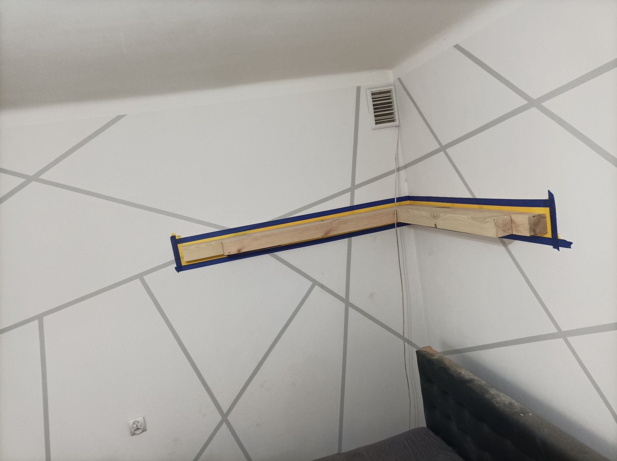 Drewniana konstrukcja przymocowana do ściany z geometrycznym wzorem za pomocą niebieskiej taśmy malarskiej.