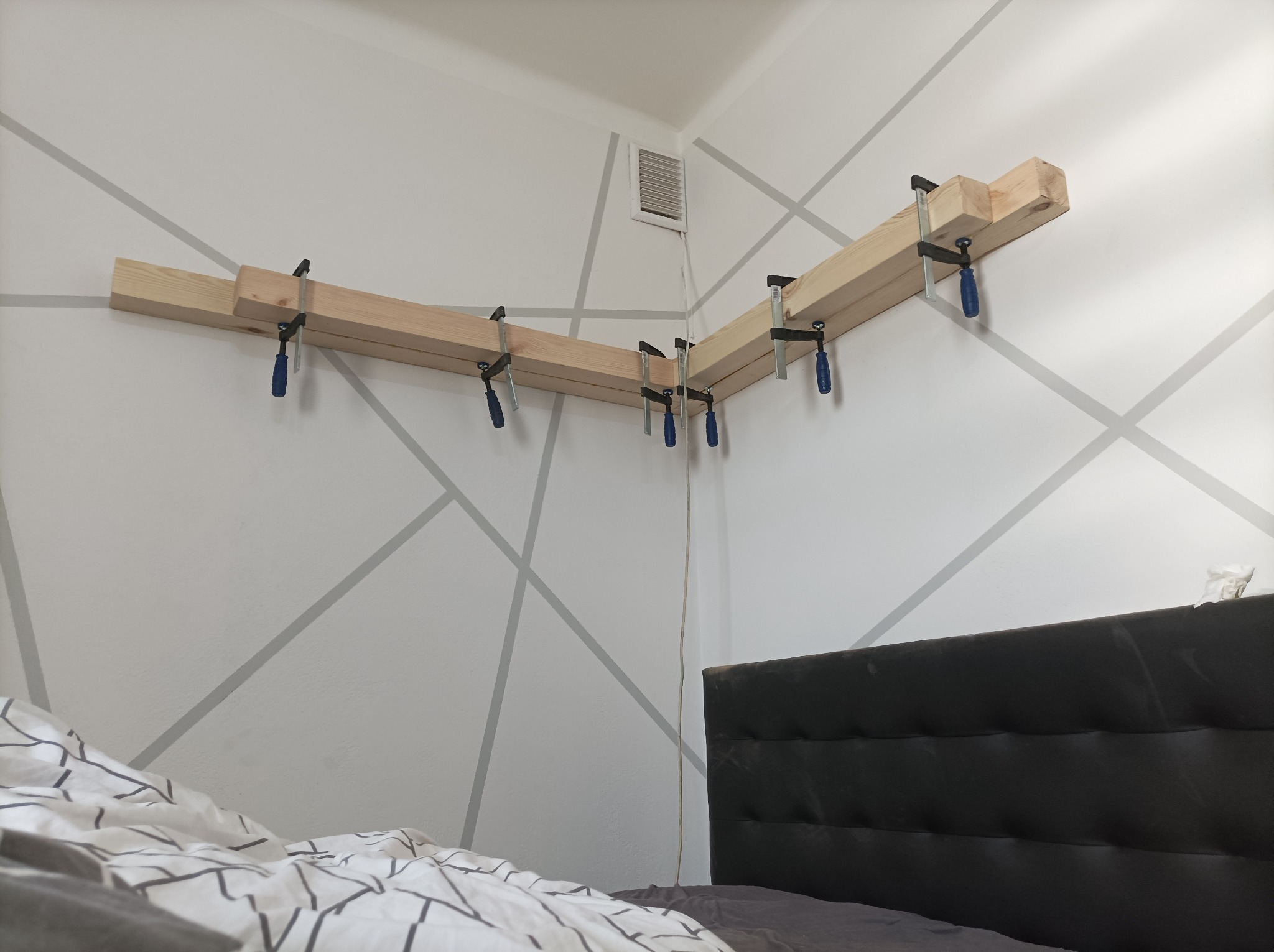 Drewniane elementy konstrukcyjne przytwierdzone do ściany za pomocą ścisków stolarskich, widoczne geometryczne wzory na ścianie i fragment sypialni.