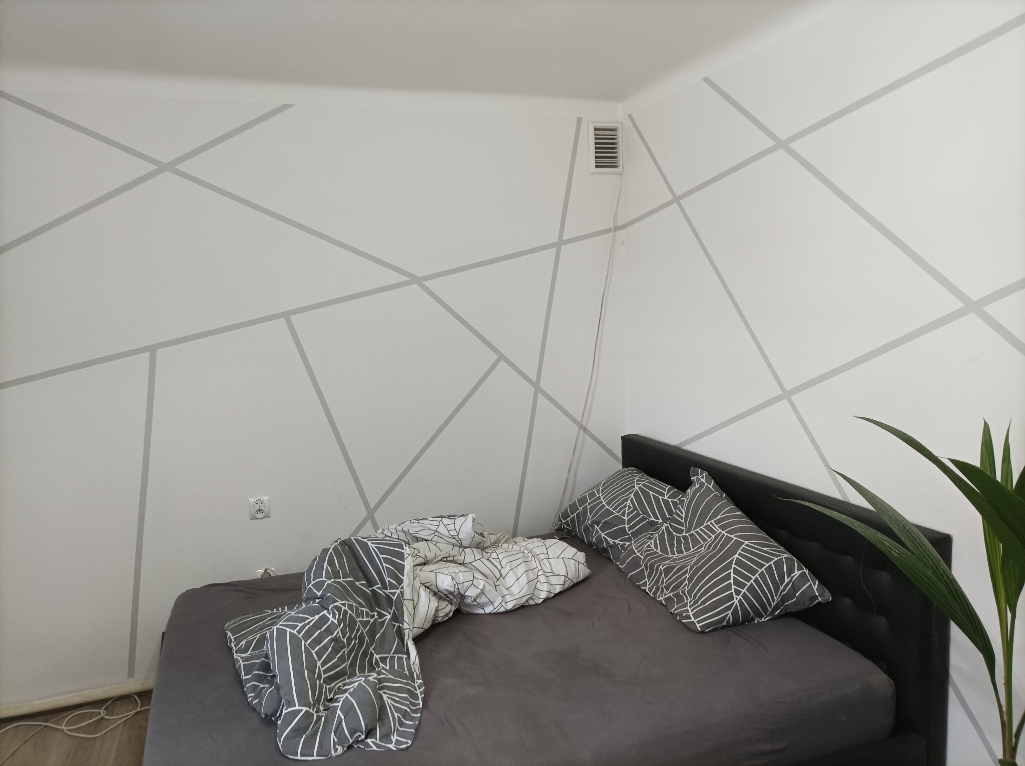 Sypialnia z łóżkiem i ścianami pomalowanymi w szare geometryczne wzory, widoczna kratka wentylacyjna i kontakt elektryczny.