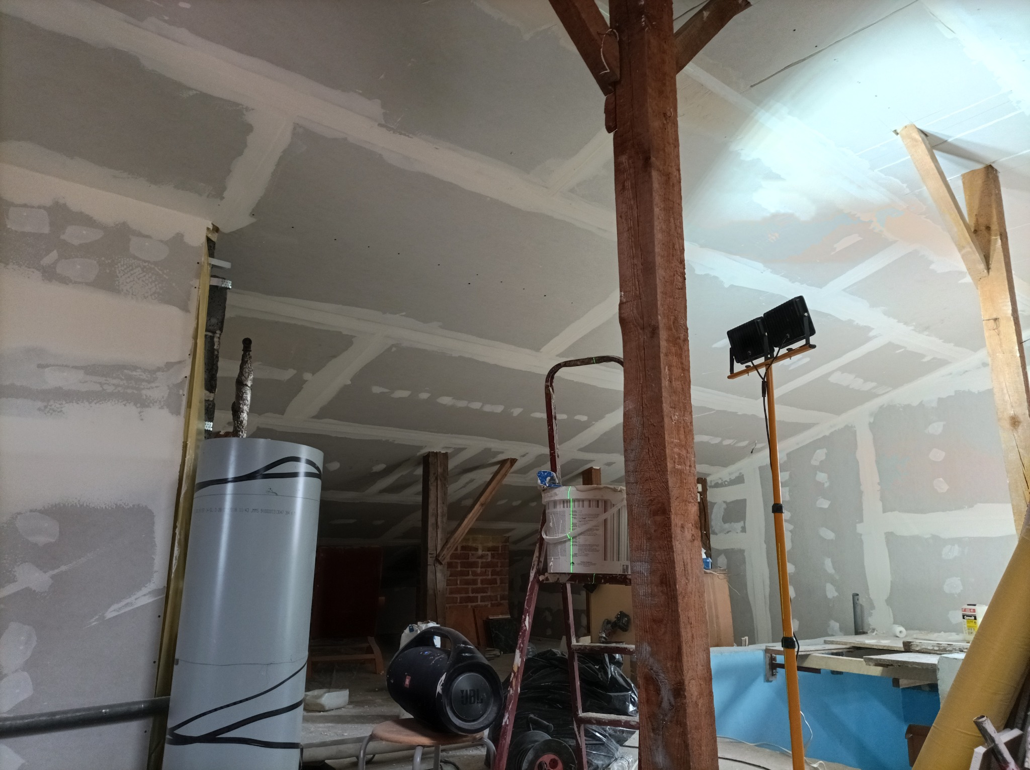 Poddasze w trakcie prac wykończeniowych: widoczne płyty gipsowo-kartonowe na suficie i ścianach, przygotowane do dalszej obróbki, drewniane słupy konstrukcyjne, drabina z wiadrem farby, lampa...