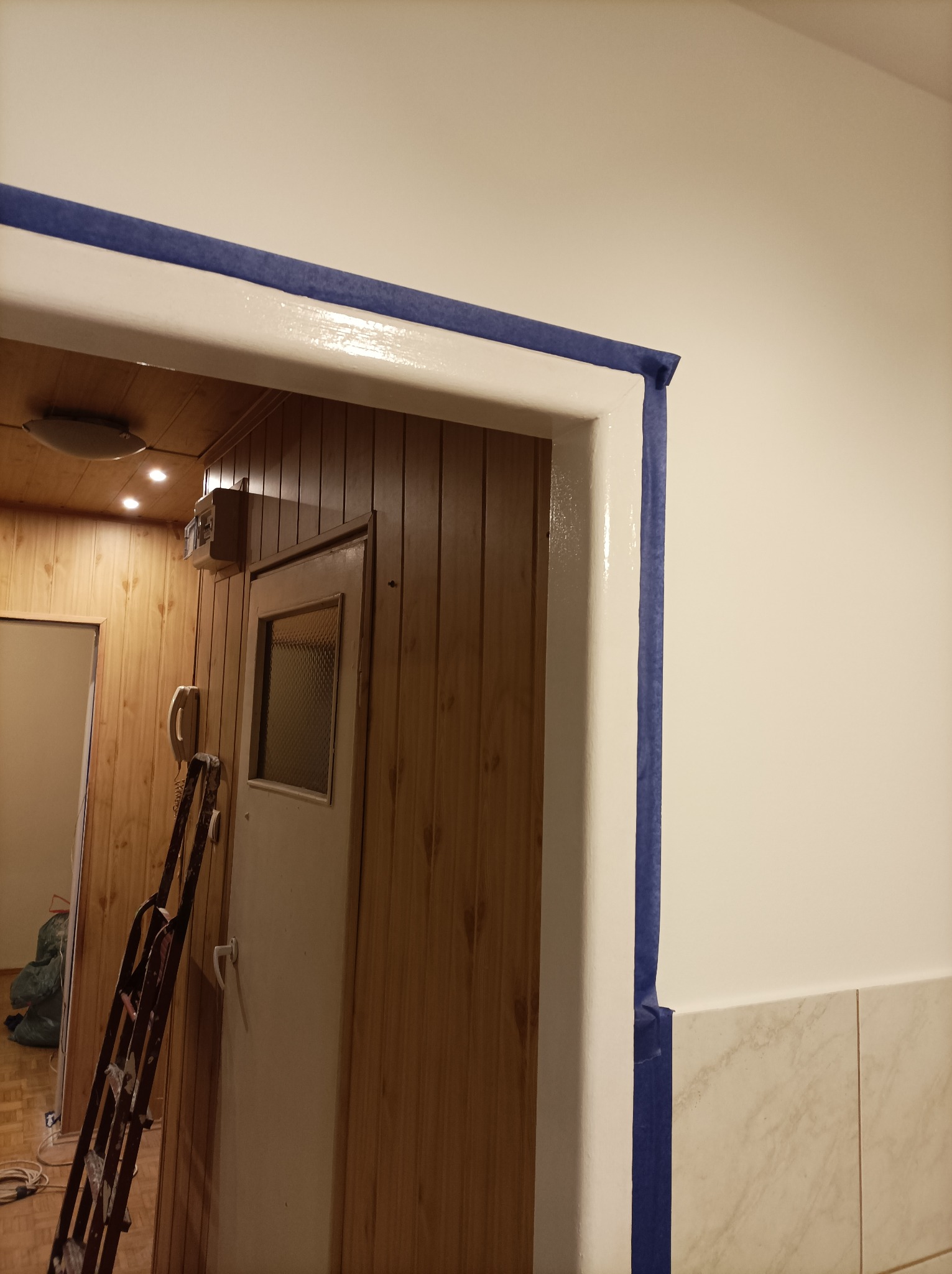 Świeżo pomalowana na biało ościeżnica zabezpieczona niebieską taśmą malarską, widoczna drabina oparta o ścianę w pomieszczeniu wykończonym boazerią.