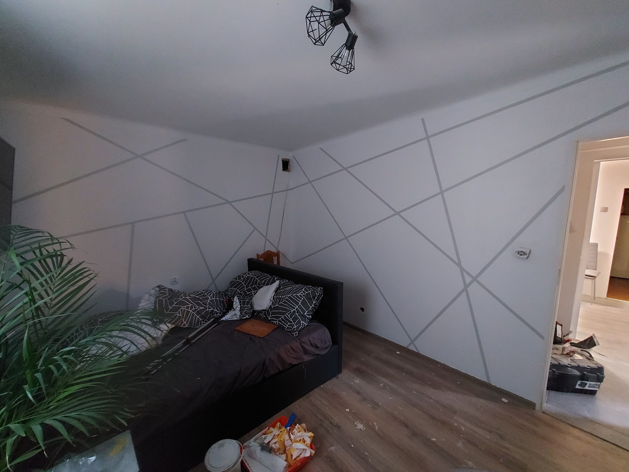 Pokój z łóżkiem i ścianami pomalowanymi w geometryczne szare linie, widoczne narzędzia malarskie i doniczka z kwiatem, etap wykończeniowy.