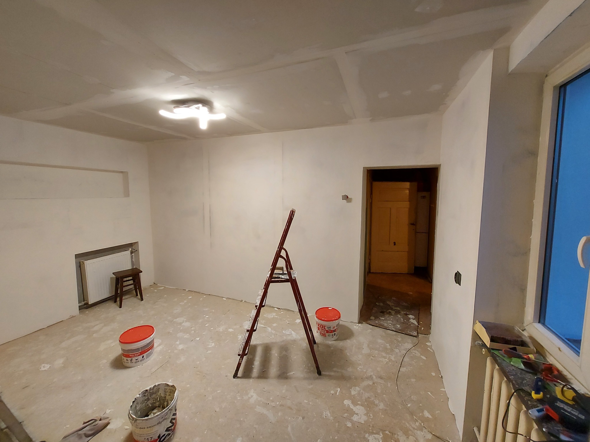 Pokój w trakcie prac wykończeniowych, ściany i sufit pokryte świeżą warstwą gładzi, widoczne narzędzia, drabina i wiadra z masą szpachlową.