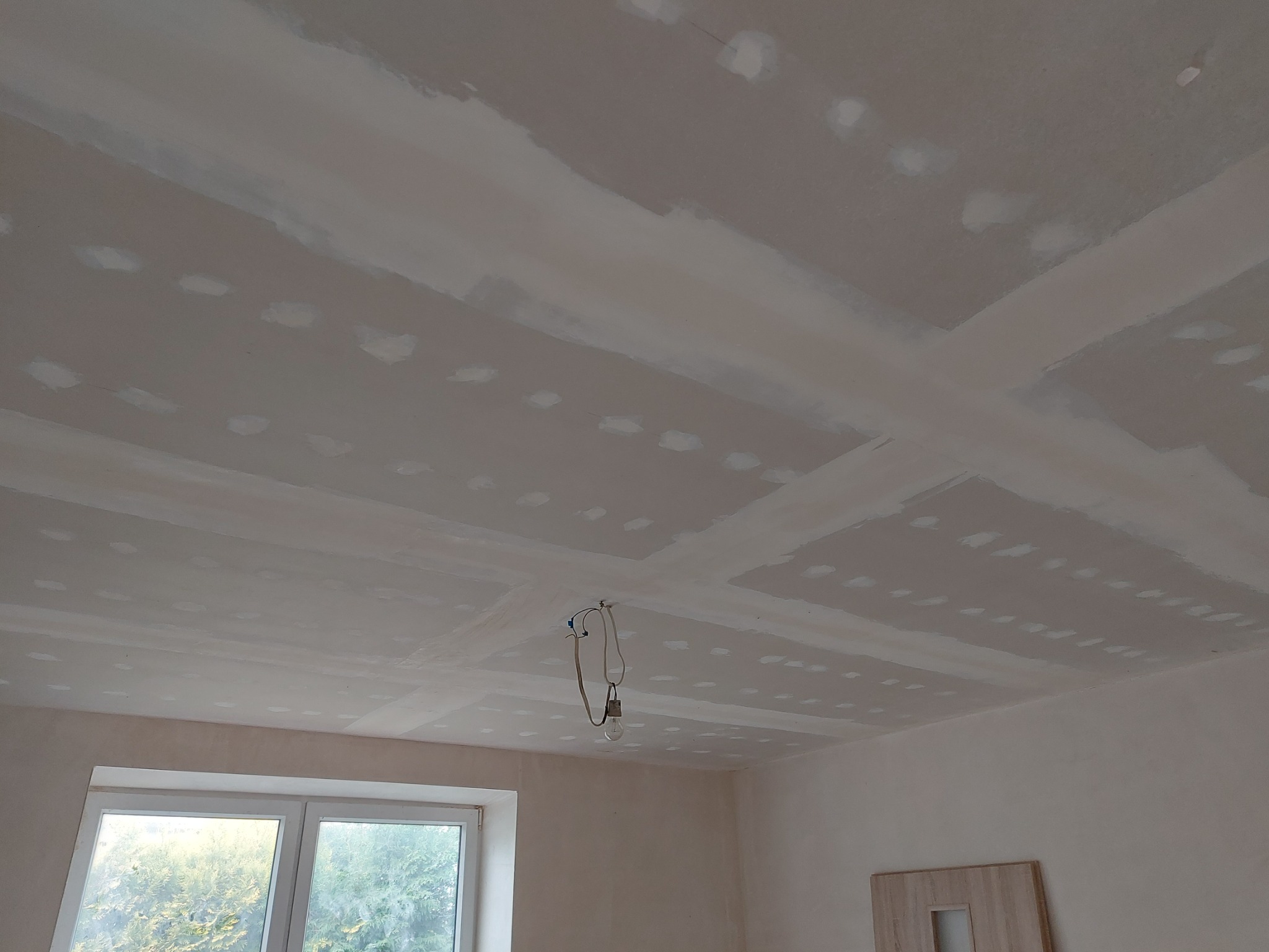 Sufit w trakcie prac wykończeniowych: widoczne płyty gipsowo-kartonowe ze świeżo nałożonymi łatami, przygotowane do dalszej obróbki, wisząca żarówka na kablu, okno w tle.