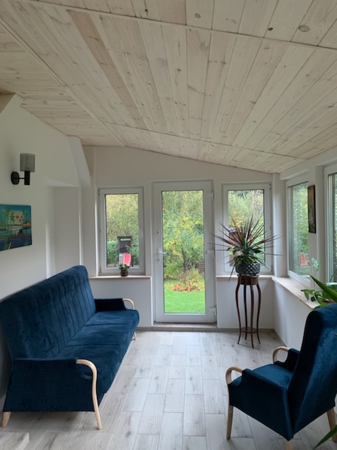 Jasne wnętrze z drewnianym sufitem z desek, błękitna sofa i fotel, widok na ogród przez okna.