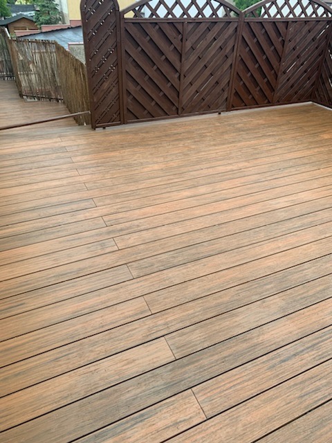 Drewniany taras z brązową kratownicą w tle, widok z góry pod lekkim kątem, deski ułożone poziomo.