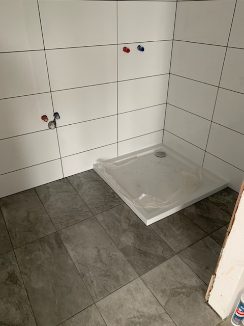 Montaż brodzika w narożnej kabinie prysznicowej wyłożonej białymi, kwadratowymi płytkami; widoczne przyłącza wodne oraz szare płytki podłogowe imitujące kamień.