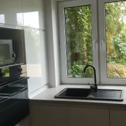 Nowoczesna kuchnia z czarnym zlewem granitowym i baterią przy oknie z widokiem na zieleń, minimalistyczne białe szafki.