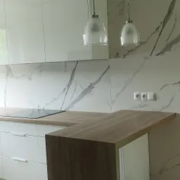 Nowoczesna kuchnia z białymi, błyszczącymi szafkami, blatem imitującym drewno i marmurowym backsplashiem. Dwie szklane lampy wiszące nad blatem. Widoczne gniazdka elektryczne na ścianie.