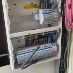 Otwarta biała skrzynka rozdzielcza z bezpiecznikami i okablowaniem w kolorach niebieskim, brązowym i żółto-zielonym, z naklejką ostrzegawczą o luźnych połączeniach, widok z bliska na instalację...
