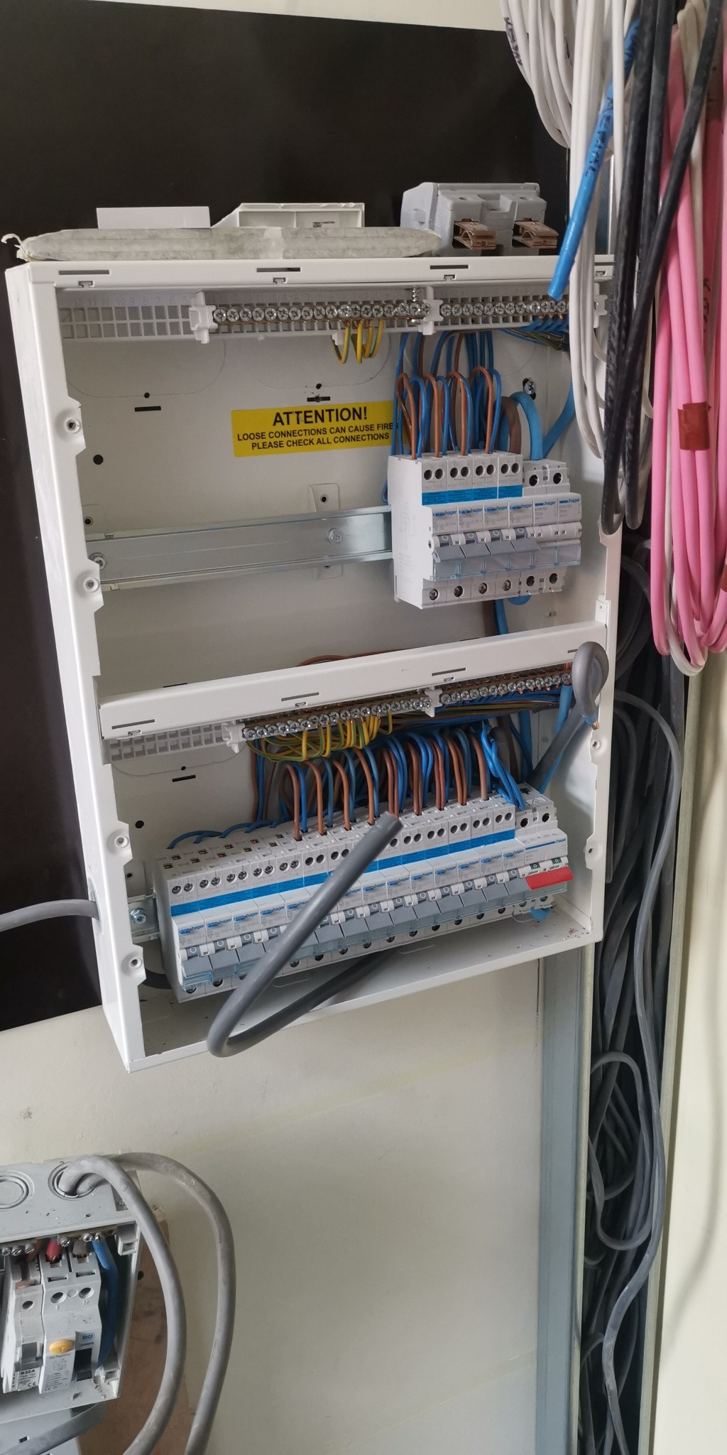 Otwarta biała skrzynka rozdzielcza z bezpiecznikami i okablowaniem w kolorach niebieskim, brązowym i żółto-zielonym, z naklejką ostrzegawczą o luźnych połączeniach, widok z bliska na instalację...