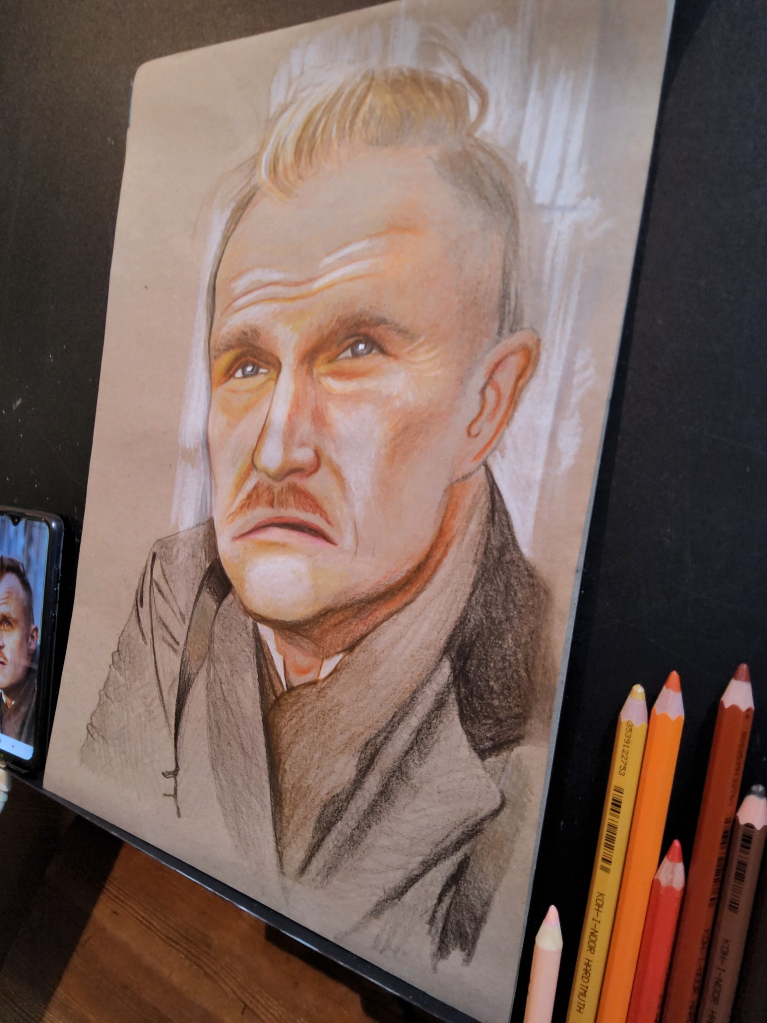 Portret mężczyzny z wąsami narysowany kredkami na brązowym papierze, obok leżą kolorowe kredki, a na telefonie wyświetla się zdjęcie tego samego mężczyzny.