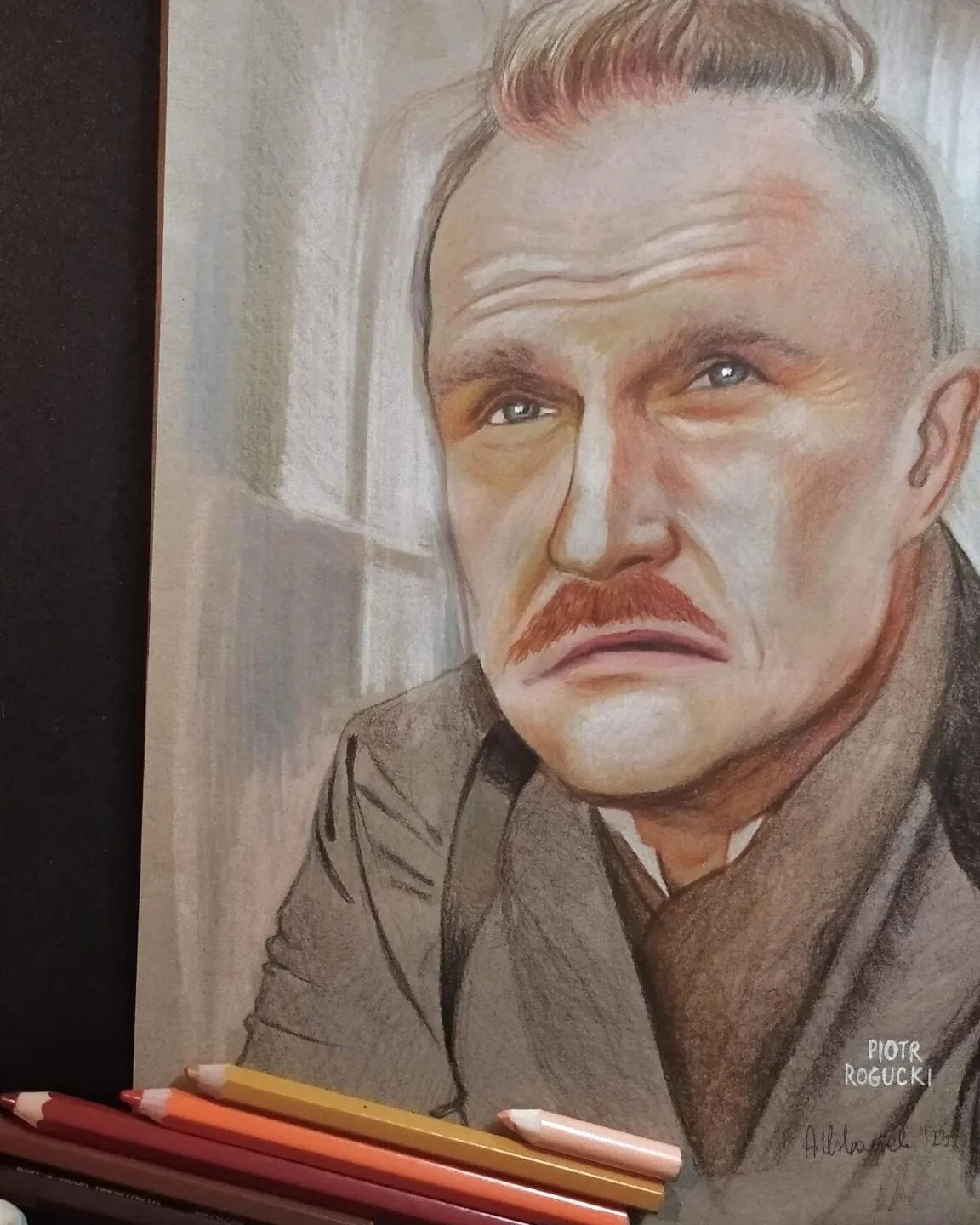 Portret mężczyzny z wąsami narysowany kredkami, widoczne kredki w odcieniach brązu i czerwieni w dolnej części kadru, podpis autora w prawym dolnym rogu.
